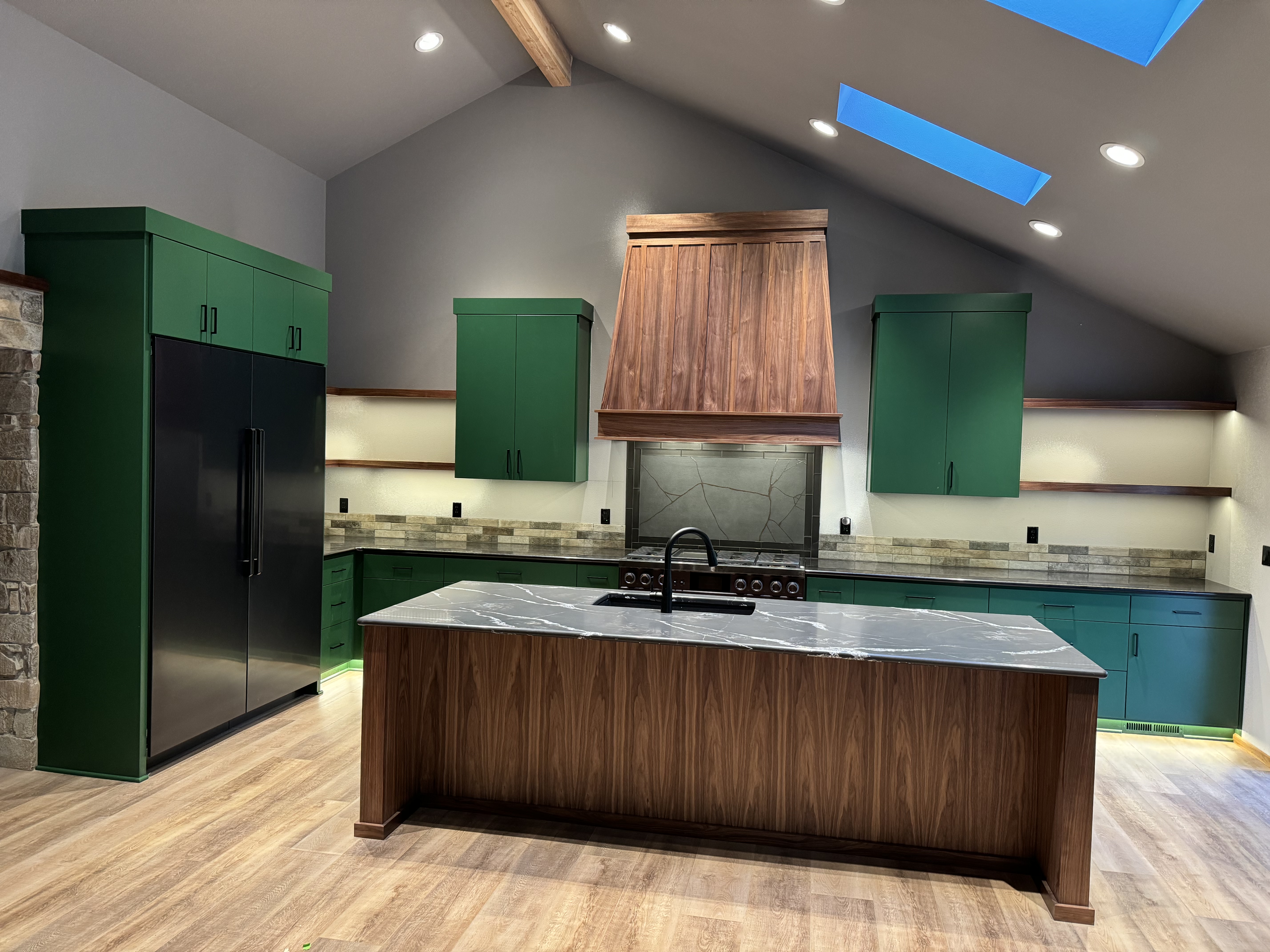 Full kitchen view showing green cabinetry, walnut island with stone countertop, and custom walnut range hood under vaulted ceiling with skylight