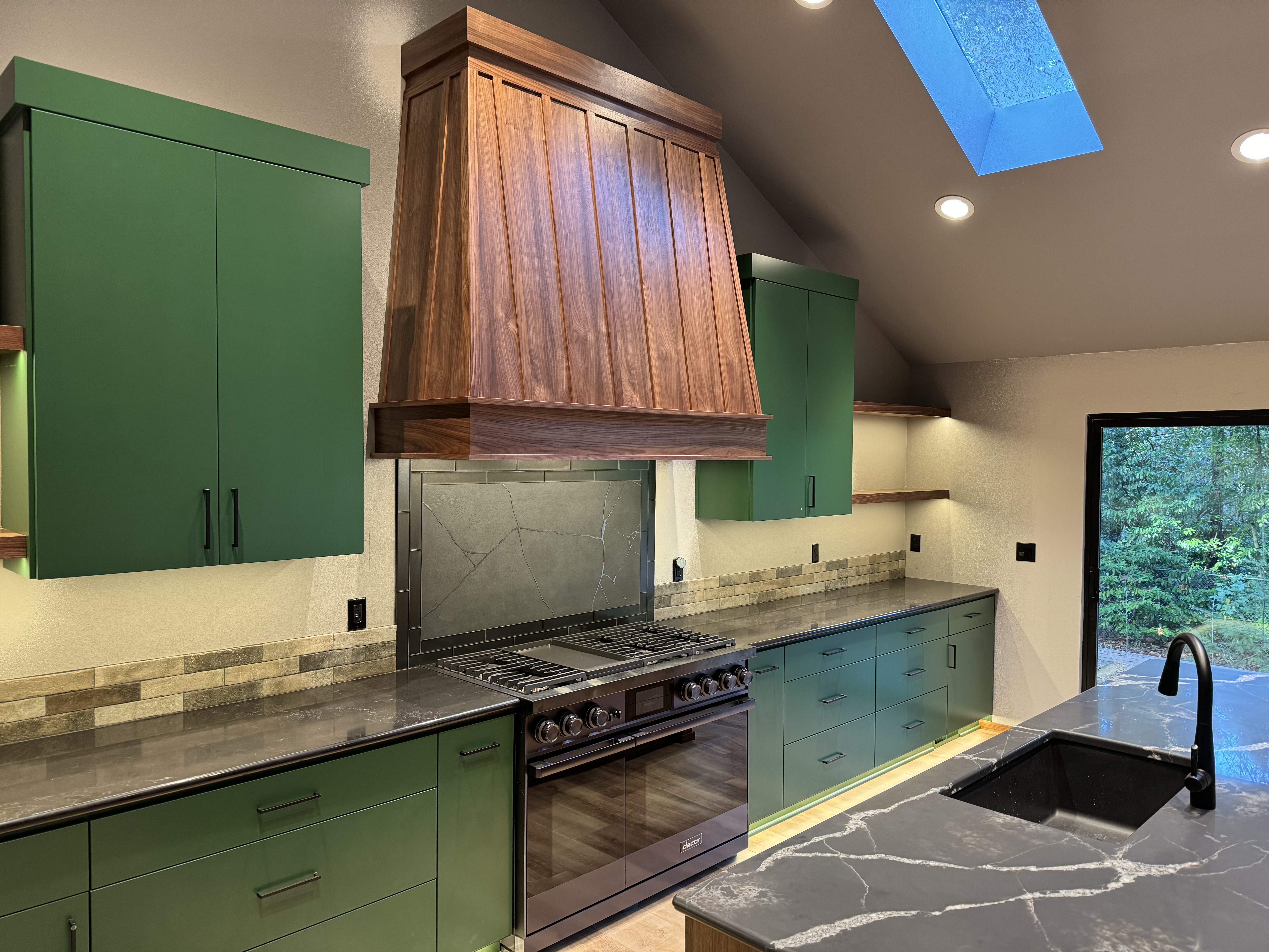 Close-up of green lower cabinets, custom walnut range hood, and dark stone slab backsplash over professional range