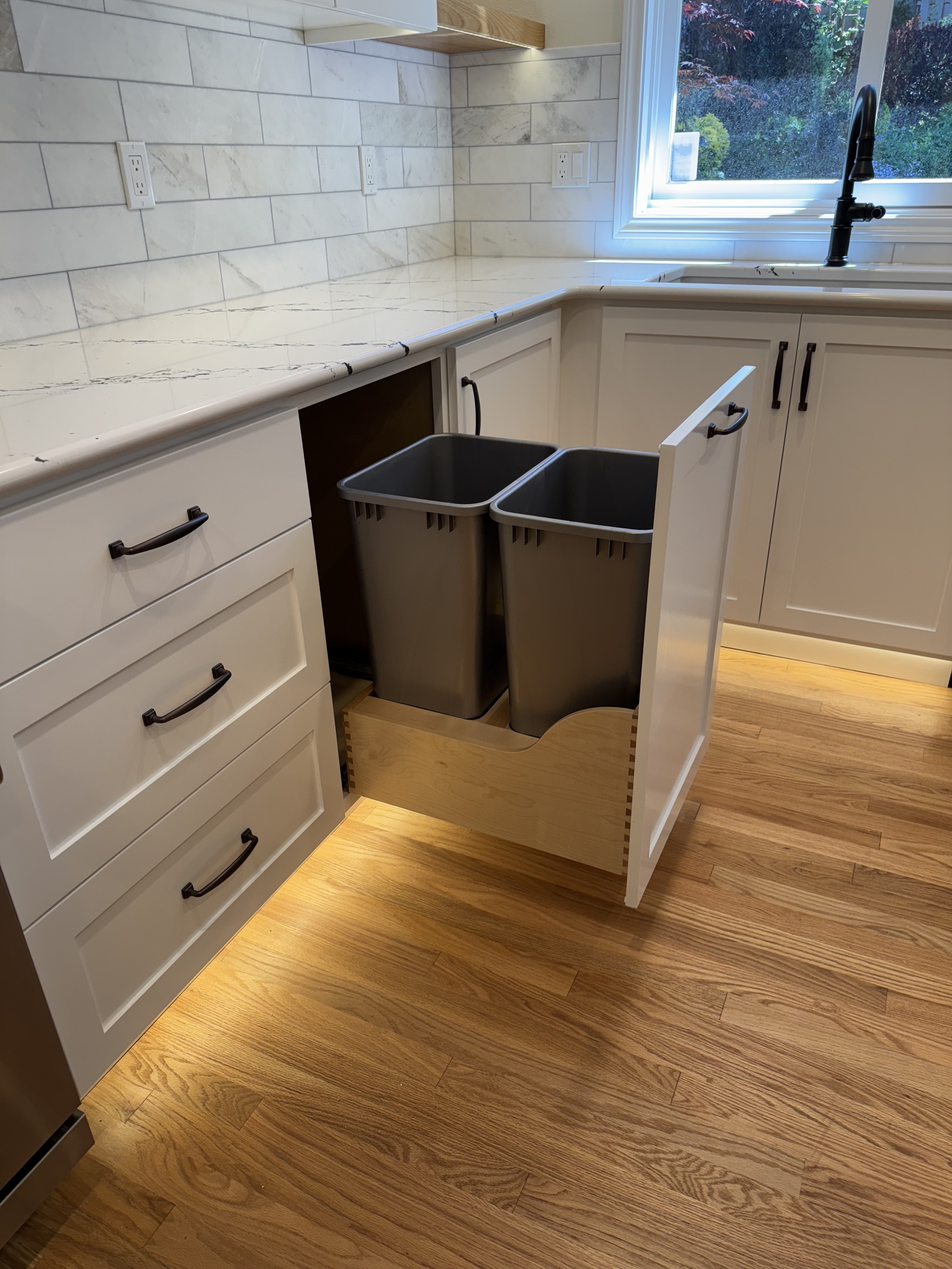 Pull-out trash and recycling cabinet with two concealed bins in a dovetailed wood drawer box behind a white shaker door