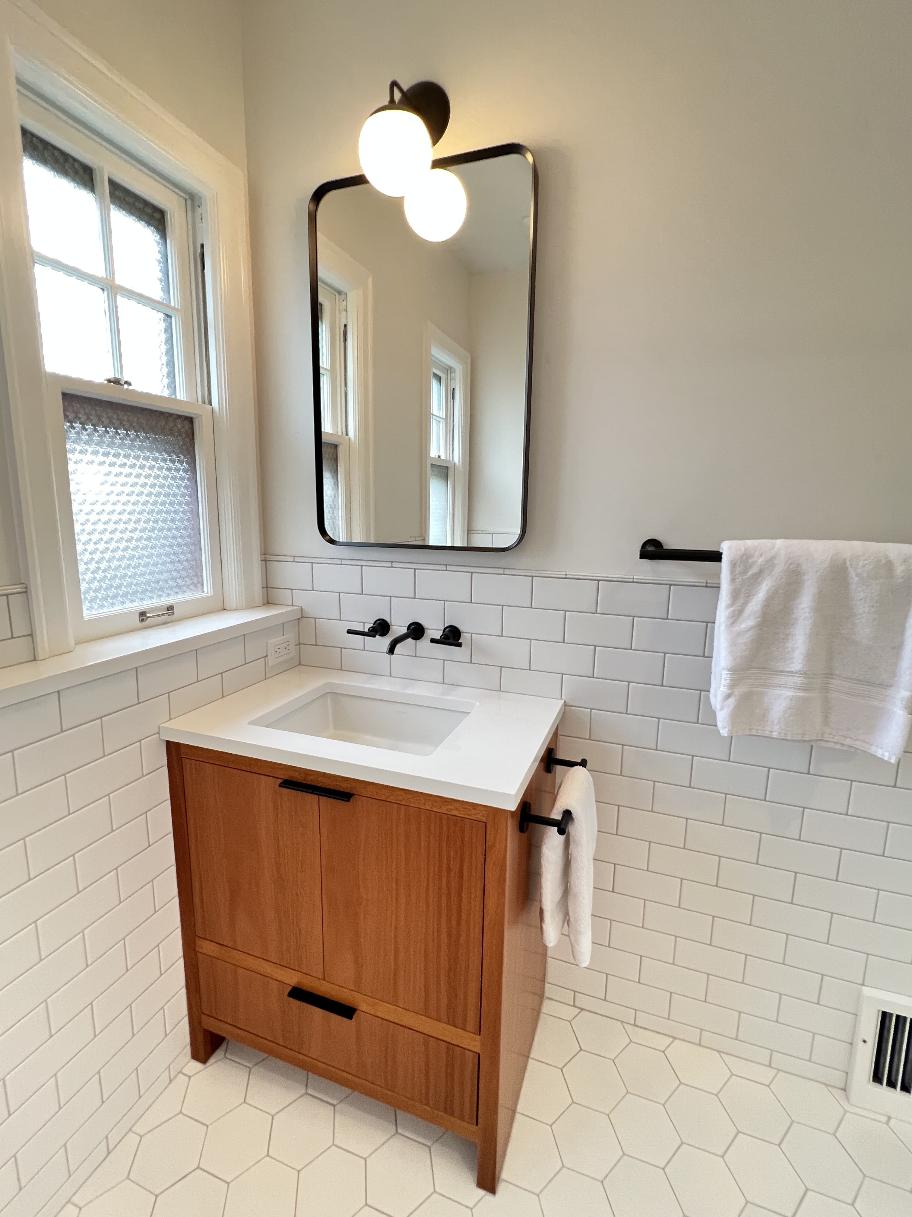 Custom warm-wood single vanity with sliding cabinet doors and matte black hardware, white subway and hex tile bathroom