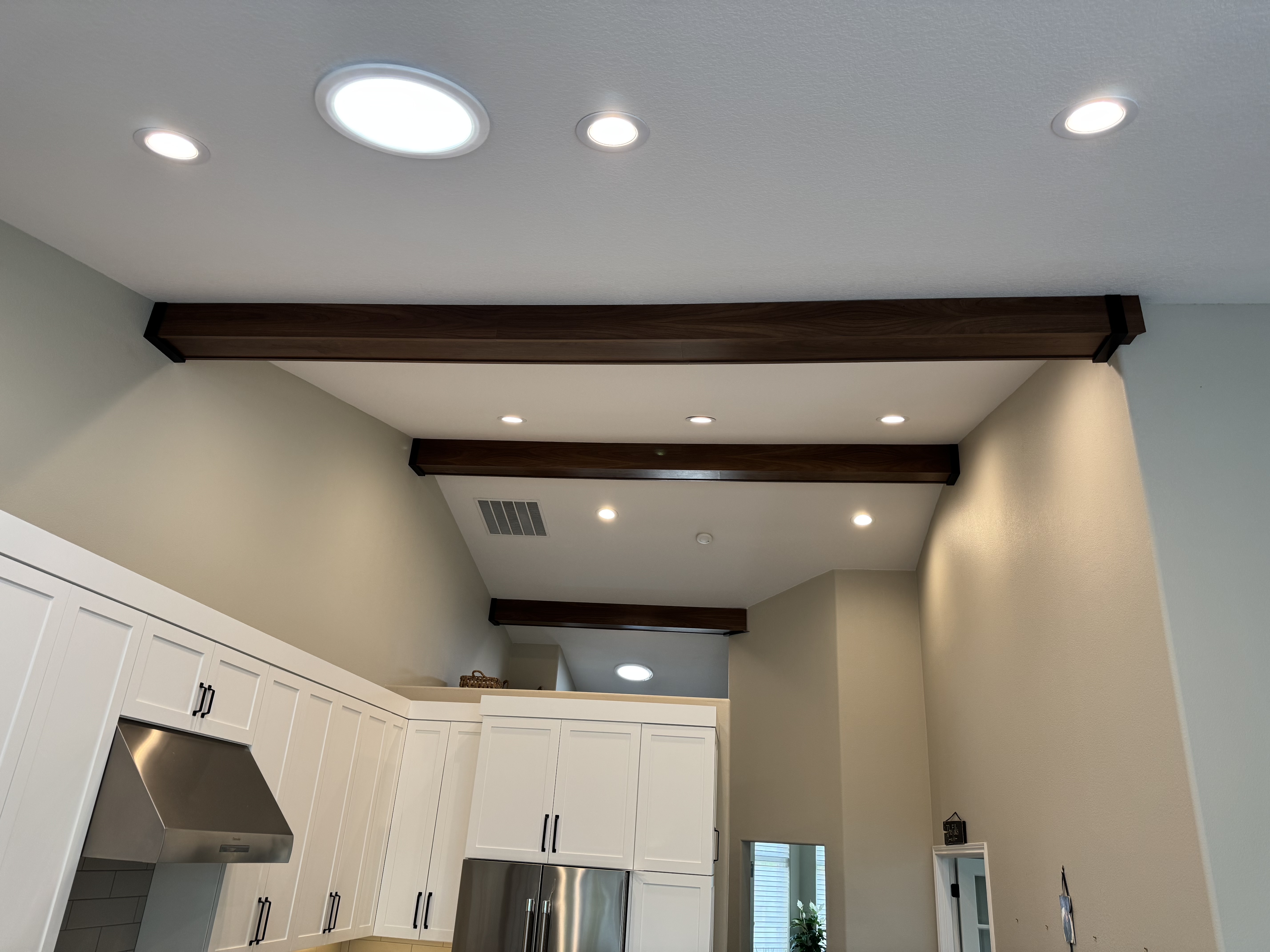 Dark walnut faux ceiling beams with recessed lighting installed over a white kitchen