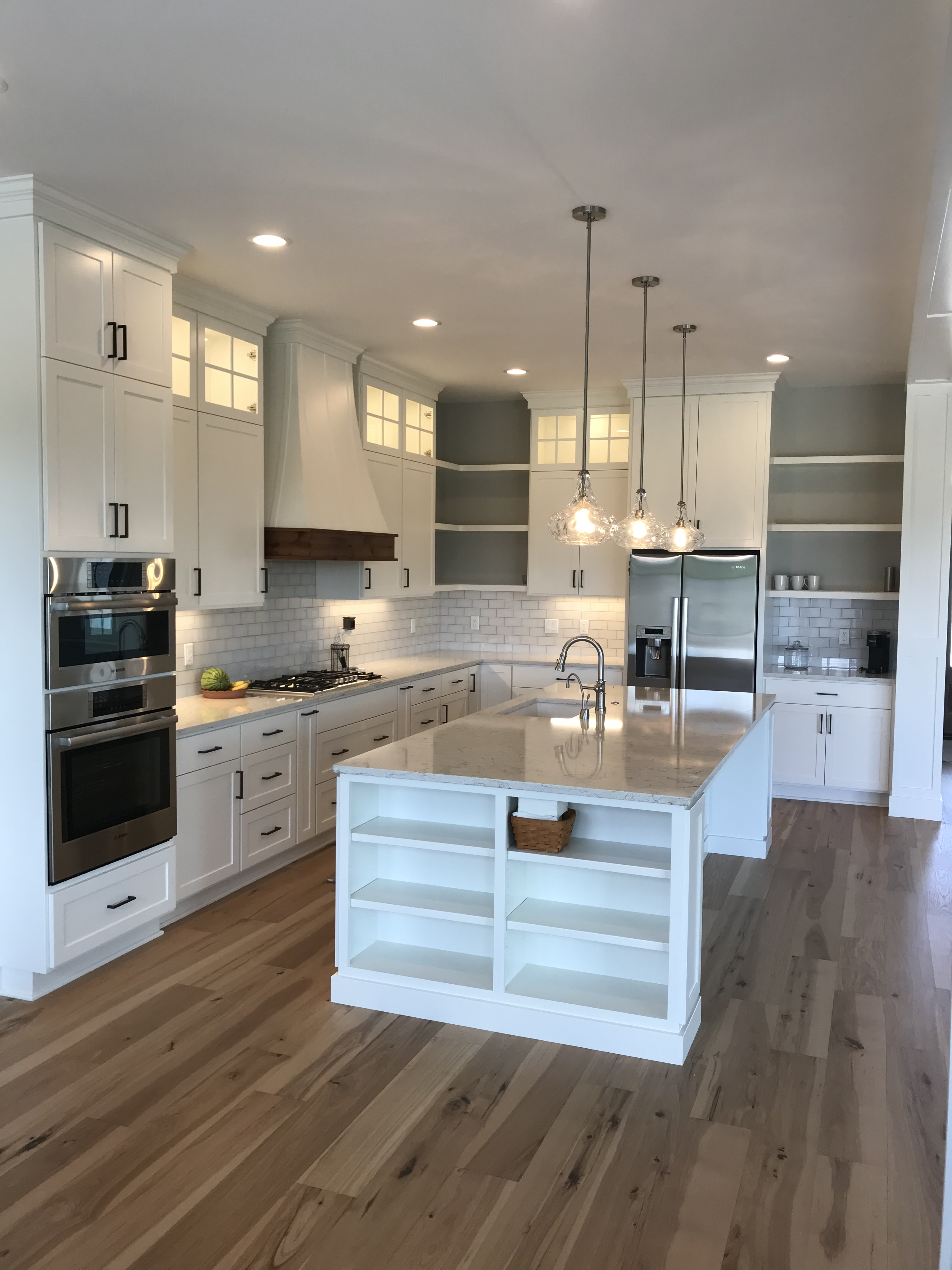 Wide view of classic white kitchen with glass-front uppers, open shelf island, pendant lights, subway tile backsplash, and hardwood floors