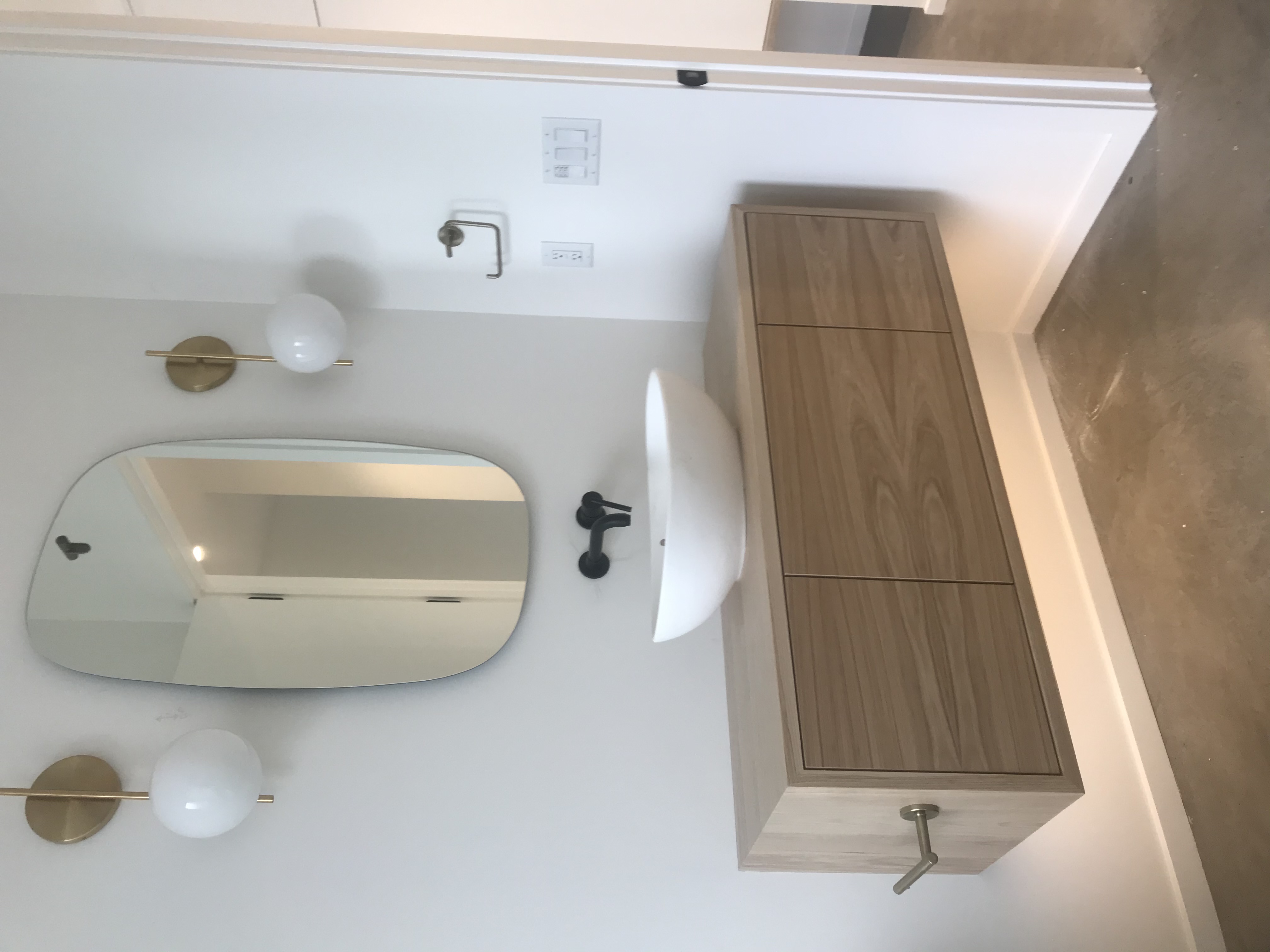 Natural wood floating single vanity with white vessel sink, black faucet, brass globe sconces, and rounded mirror