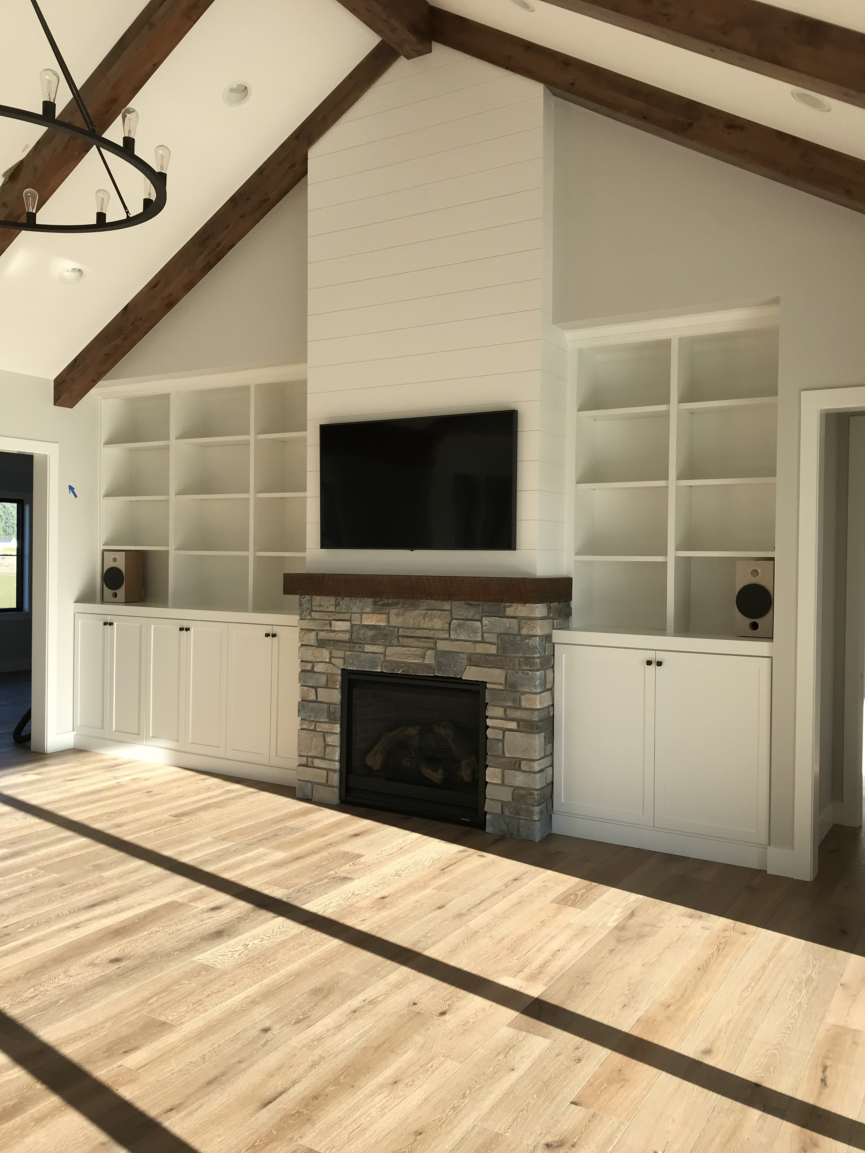 White built-in flanking a stone fireplace with TV above wood mantel, open cubbies, and shiplap accent wall under vaulted ceiling
