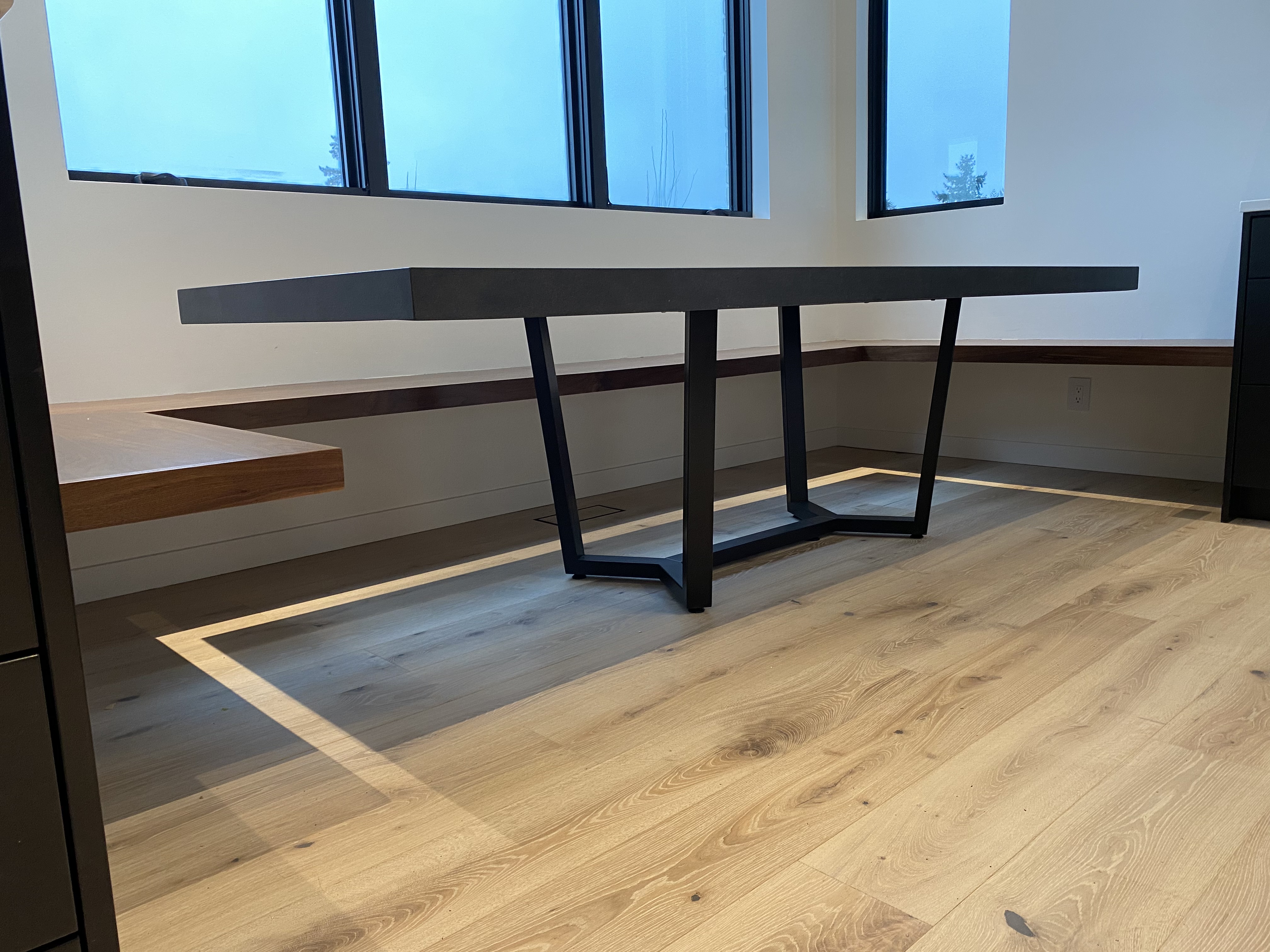 Finished walnut floating bench seat wrapping a window corner with a custom dark matte dining table on a geometric black steel base