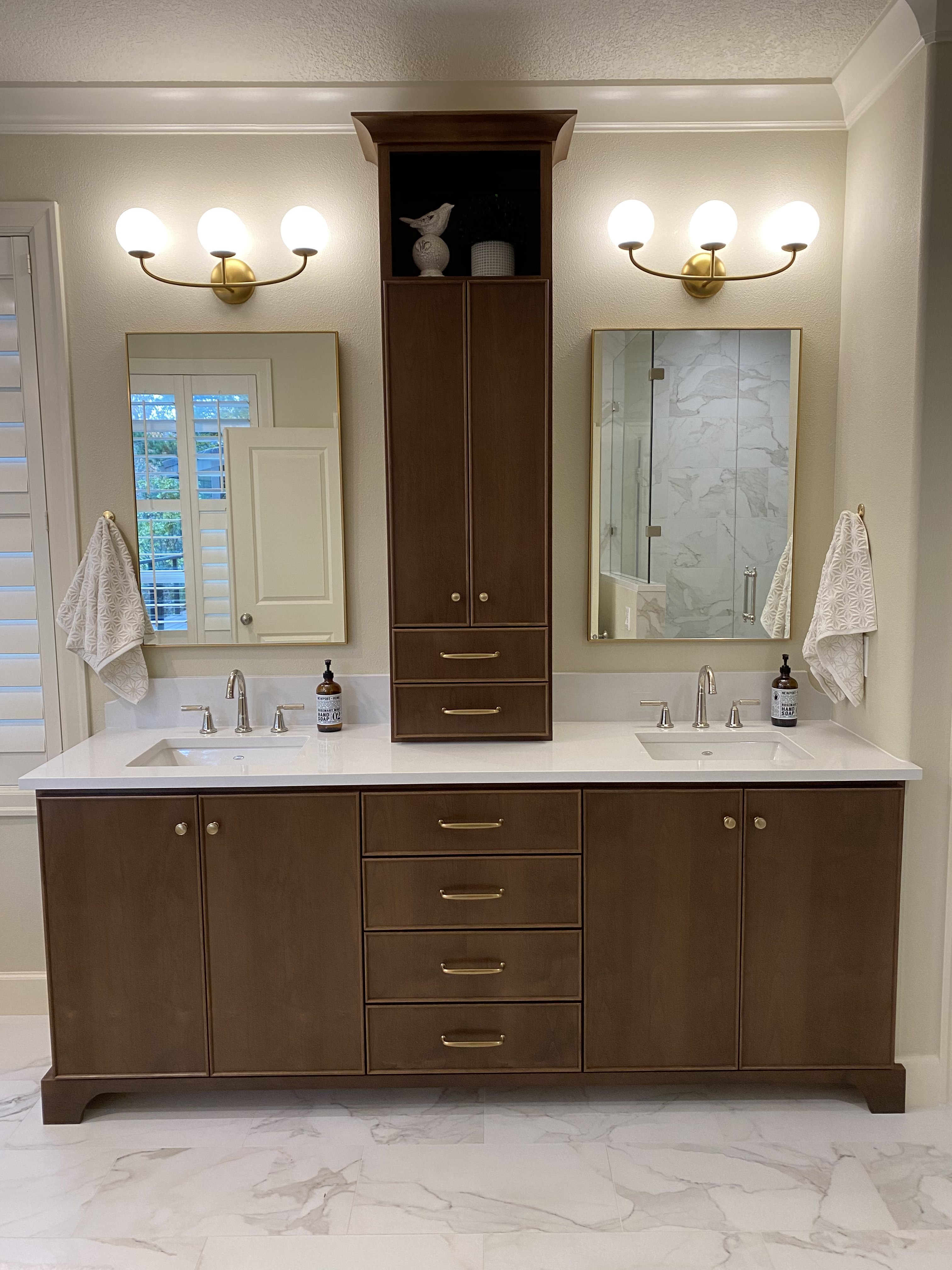 His-and-hers walnut bathroom vanities with brass hardware, medicine cabinets, tall linen tower, and marble-look tile surround