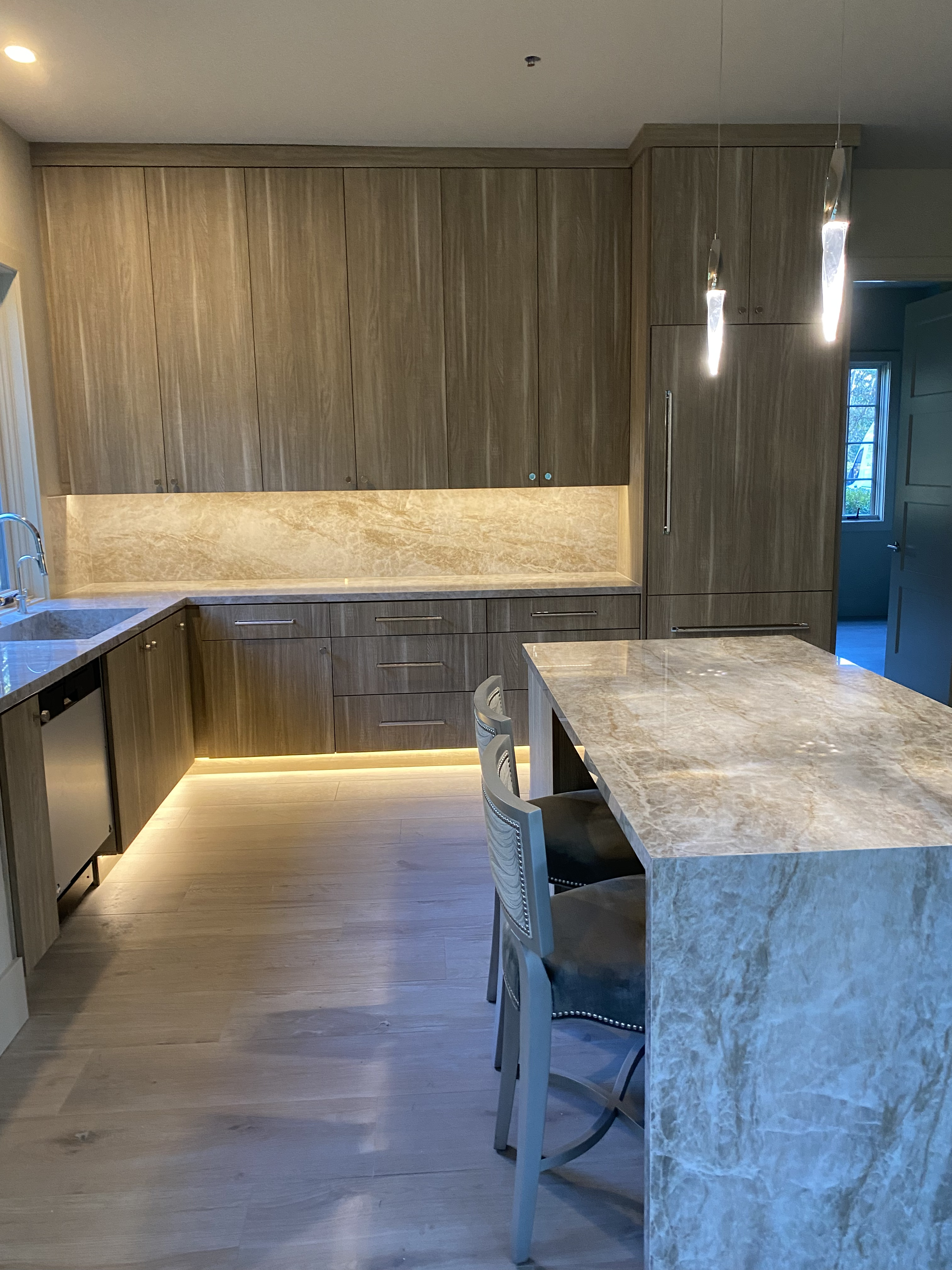 Wood grain slab-front kitchen with floor-to-ceiling uppers, stone waterfall island, pendant lights, and LED toe-kick lighting